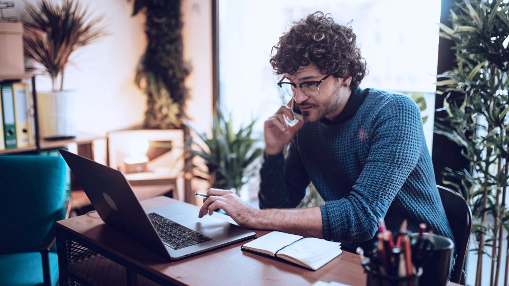 Foto de um homem sentado a uma mesa com computador, enquanto fala no celular, trabalhando em casa.
